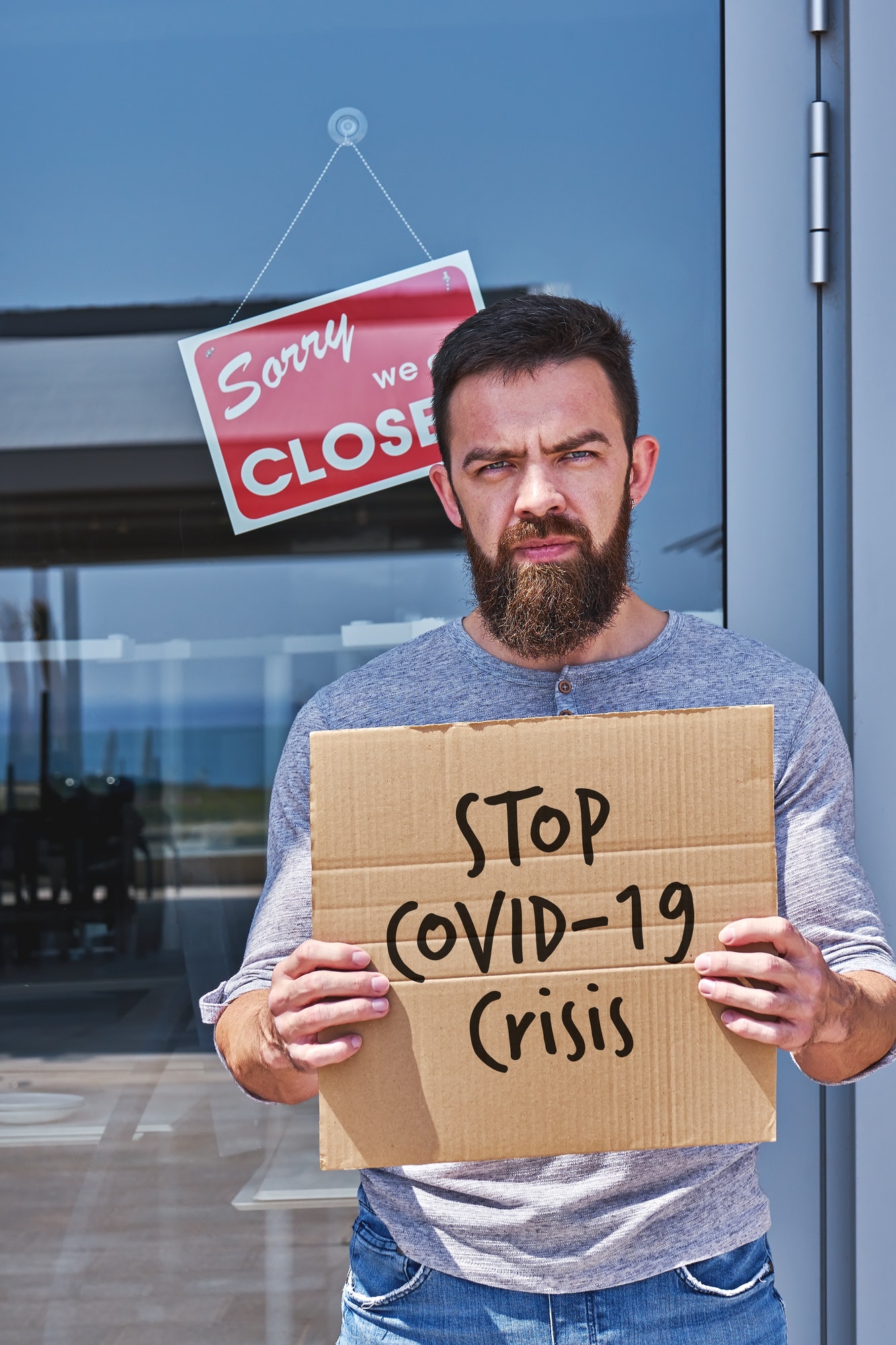 Unemployed guy with sign STOP COVID-19 CRISIS near closed door office or cafe